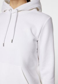 Vit sweatshirt med kängurutaske, dragsko i huvan och subtil logotyp. Tillverkad av mjuk, texturerad tyg med metalliska detaljer på dragskoarna.