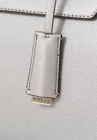 Borsa sintetica argento con texture, caratterizzata da un grande tag GUESS, design elegante e dettagli di cucitura discreti sulla tracolla.