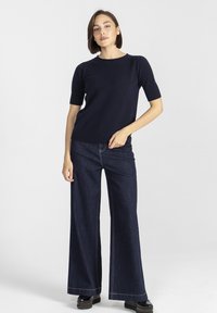 Kobieta stojąca na białym tle, ubrana w granatowy sweter z krótkim rękawem, ciemnogranatowe dżinsy z szerokimi nogawkami oraz czarne buty na platformie.