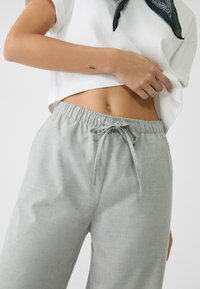 Kobieta w białej wyciętej bluzce, szarych spodniach z troczkami i czarną chustą na szyi, ukazująca swoje brzuch i dłoń.