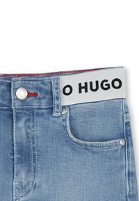 Jeans de mezclilla azul claro con una cinta ancha que lleva escrito "O HUGO" en negro, un botón plateado y detalles en costura roja.