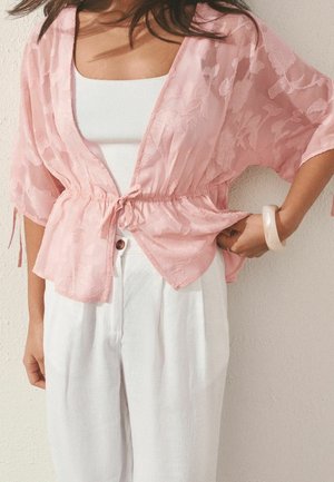 Femme portant un chemisier cache-cœur texturé à fleurs rose clair par-dessus un débardeur blanc et un pantalon blanc ample taille haute, avec un bracelet pâle à son poignet droit.