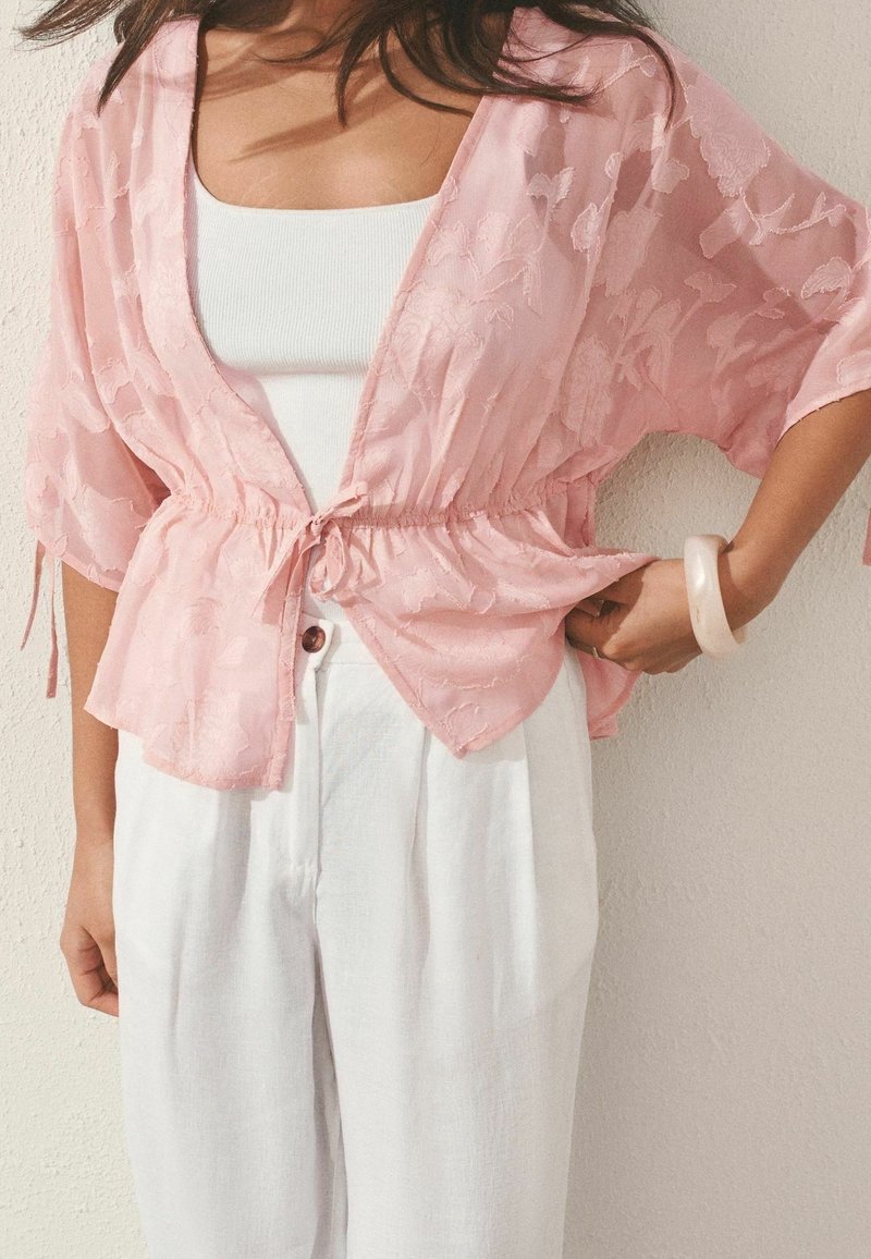 Femme portant un chemisier cache-cœur texturé à fleurs rose clair par-dessus un débardeur blanc et un pantalon blanc ample taille haute, avec un bracelet pâle à son poignet droit.