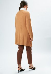 Kamelfarvet oversized strikket cardigan med kabelstrik-detaljer, parret med brune slim-fit bukser og sorte slingback hæle med udsmykning.