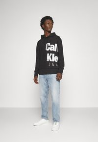 Felpa nera con grande testo bianco "Calvin Klein JEAN", abbinata a jeans azzurri e sneakers bianche. Texture liscia, vestibilità casual.