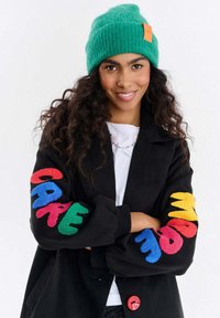 Kobieta z długimi kręconymi włosami, ubrana w zieloną dzianinową czapkę i czarną kurtkę z kolorowymi naszywkami "CARE" i "MADE" na rękawach, uśmiechająca się ze skrzyżowanymi ramionami.