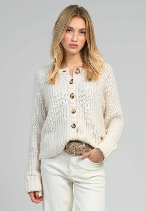 Cardigan en tricot de couleur crème avec une fermeture à boutons, comportant cinq grands boutons et des poignets côtelés. Assorti à un pantalon blanc et une ceinture tressée.
