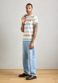Mann med tatoveringer iført stripete t-skjorte, lyseblå jeans og mørke loafers, holder en jakke over skulderen mot en ubrutt bakgrunn.