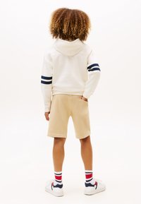 Felpa crema con strisce blu navy, pantaloni corti beige e sneakers bianche. La vista posteriore mostra capelli ricci e calzini con strisce rosse, bianche e blu.