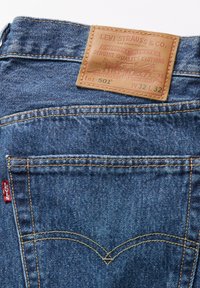 Jeans in denim blu con una toppa in pelle marrone sulla vita, caratterizzati da cuciture gialle e un design della tasca posteriore curvo.