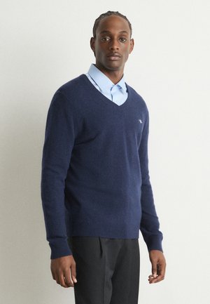 Granatowy sweter w serek wykonany z miękkiej tkaniny, w połączeniu z jasnoniebieską koszulą z kołnierzykiem, z prążkowanymi mankietami i dolnym brzegiem. Model stoi na jednolitym tle.