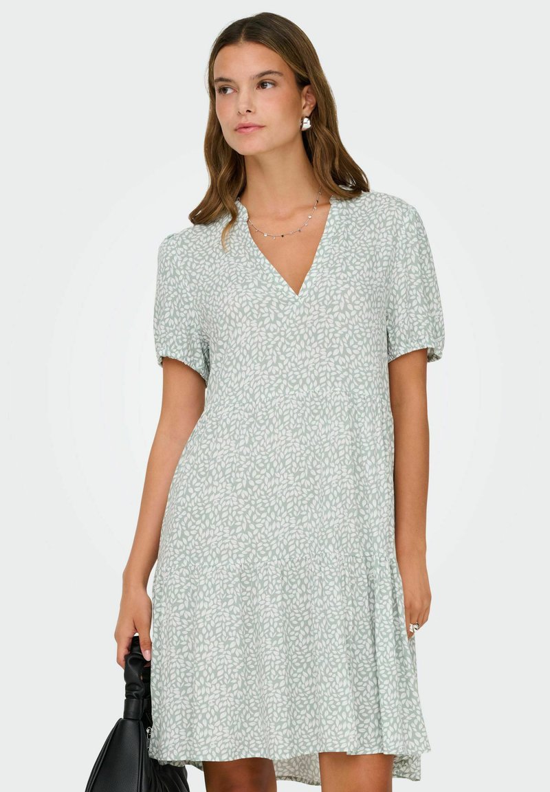 Femme portant une robe à manches courtes à motifs de feuilles vert clair, tenant un sac à main noir, regardant légèrement sur le côté.