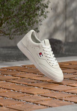 Baskets basses blanches avec lacets et un logo rectangulaire rouge, blanc et bleu flottant au-dessus d'un sol en dalles de bois en extérieur.