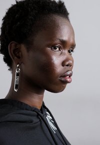 Sølvøredobber med geometrisk design, hengende fra en krok. Personen har kort, teksturert hår og har på seg en svart hettegenser.