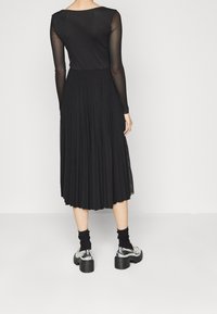 Robe noire plissée avec des manches longues en dentelle, un col rond et une coupe jusqu'aux chevilles, associée à des chaussures à talons épais en argent et des chaussettes noires.