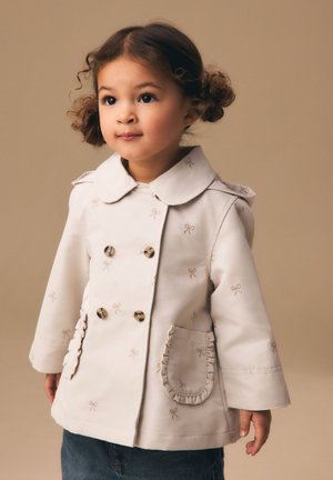 Niño pequeño con cabello rizado que lleva un abrigo cruzado de color claro con patrones de lazos y bolsillos con borde fruncido, de pie sobre un fondo beige.