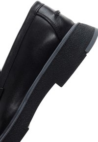 Zapato de cuero negro con suela de goma texturizada y un diseño minimalista. Presenta una puntera redondeada y costuras sutiles a lo largo de la parte superior.