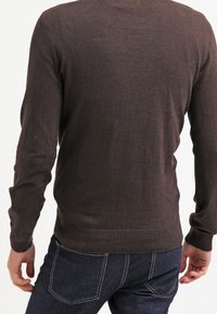 Brun stickad tröja med lång ärm, ribbad nederkant och rundad hals, tillverkad av mjukt material. Bärs över mörka denimjeans.