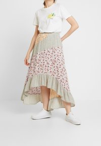 Jupe longue à volants avec des tissus floraux et à motifs dans des tons pastel, associée à un t-shirt blanc à imprimé graphique et des baskets blanches.