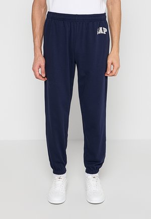 Mężczyzna noszący granatowe spodnie dresowe GAP z elastycznym pasem i mankietami, połączone z białymi trampkami i białymi skarpetkami.