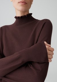 Brauner, gestrickter Pullover mit hohem, rüschendem Kragen und langen Ärmeln. Der Stoff wirkt weich mit einer glatten Textur. Die figurbetonte Passform betont die Silhouette.