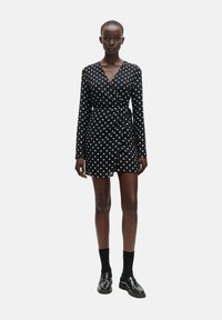 Zwarte wikkeljurk met lange mouwen, voorzien van witte stippen, een diepe V-hals en een getailleerde taille. Draagt met zwarte sokken en nette schoenen.