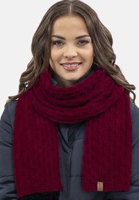 Sciarpa lavorata a maglia color bordeaux con un motivo testurizzato, appoggiata attorno al collo. Realizzata in tessuto morbido, presenta un bordo cucito e un'etichetta.