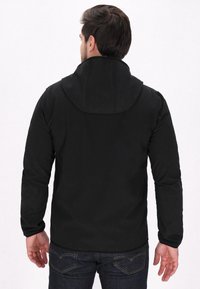 Melna kapuce žakete ar gludu tekstūru, pieguļošs dizains, garas piedurknes un izliekta apakšmala. Valkāta virs tumšiem džinsiem, izceļot minimālistisku stilu.