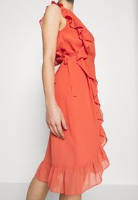 Koralowa sukienka midi z falbanką wzdłuż dekoltu i boków, gładka tkanina, dopasowana talia i rozkloszowany, falbaniasty dół.