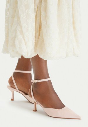 Ciemnoskóre stopy w jasnoróżowych, szpiczastych koturnach z paskami wokół kostki pod białą, kwiatową koronkową spódnicą midi.