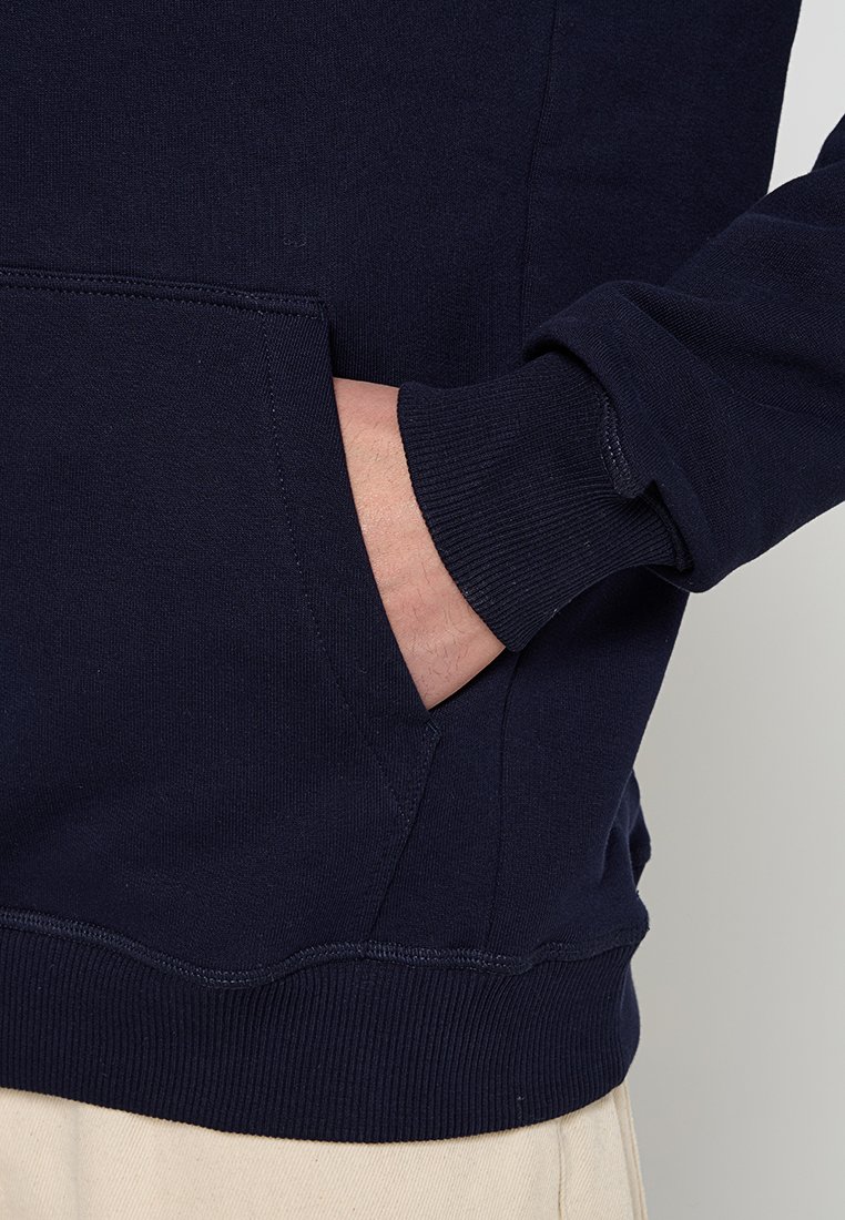 Persona che indossa una felpa blu navy con una mano parzialmente dentro la tasca frontale, pantaloni beige visibili nel margine inferiore.