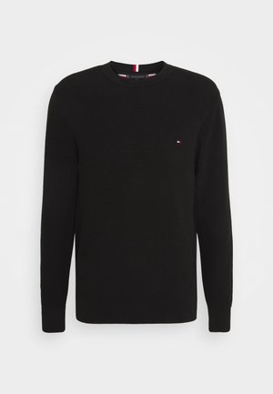 Schwarzer Pullover mit langen Ärmeln, einem strukturierten Strickmuster und einem runden Halsausschnitt. Kleines Logo auf der Brust mit roten und weißen Akzenten.