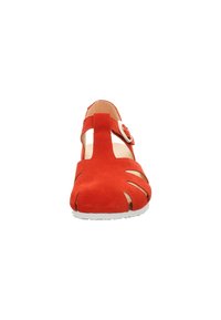 Red suede shoe with cut-out detailing, a strap with a circular metal buckle, and a white rubber sole. Front view highlights design elements.