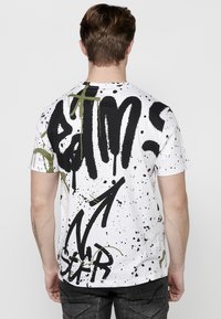 Camiseta blanca con gran texto en estilo graffiti negro y salpicaduras de pintura verde. Manga corta, cuello redondo, ajustada de forma casual.