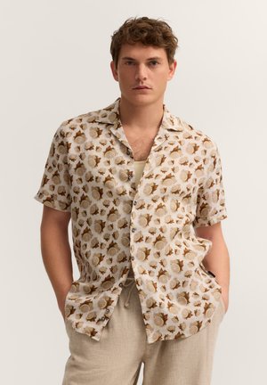 Mann trägt beige gemustertes kurzärmeliges Hemd über cremefarbenem Tanktop mit beiger Hose, steht mit Händen in den Taschen vor neutralem Hintergrund.