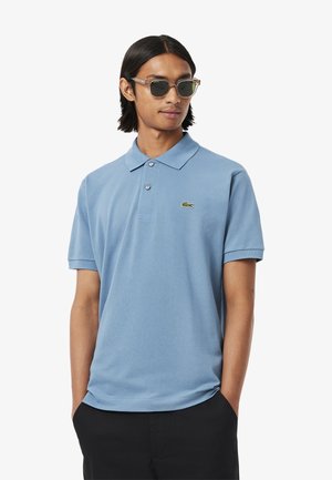 Polo shirt azzurro in tessuto morbido, con colletto, due bottoni e un piccolo logo verde sul lato sinistro del petto. Indossato con pantaloni neri.