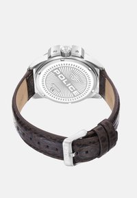 Bruin leren horlogeband met textuurdetail, zilveren metalen kast, zichtbaar logo op witte wijzerplaat en prominente kroon op de positie van 2 uur.