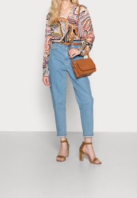 Blusa a maniche lunghe colorata e a fantasia, jeans a vita alta di colore blu chiaro, sandali con tacco marroni e una piccola borsa marrone.