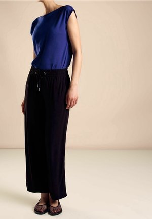 Kobieta w niebieskim topie bez rękawów, luźnych czarnych spodniach z wiązaniem i czarnych siateczkowych balerinach, stojąca na beżowym tle.