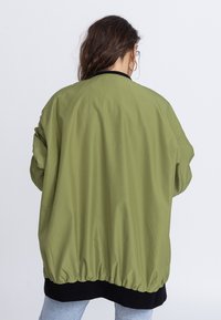 Veste bomber verte olive avec un ourlet noir côtelé, des poignets élastiques et une texture lisse, légèrement brillante. Coupe loose avec des épaules tombantes.