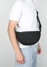 Uomo che indossa una polo bianca e jeans azzurri chiari, porta una borsa a tracolla nera appesa alla vita.