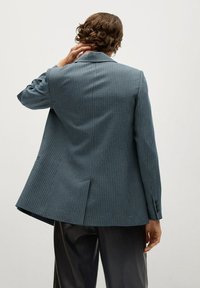 Blazer à carreaux bleus avec des épaules structurées, une fente au dos, et des poignets à bouton, associé à un pantalon sombre ajusté. Tissu texturé.
