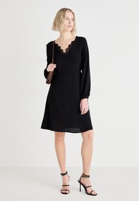 Μαύρο φόρεμα με μακριά μανίκια, V-λαιμόκοψη με δαντελένια τελειώματα, φούστα σε Α γραμμή και λεία υφή υφάσματος. Συνδυάζεται με μαύρα ψηλοτάκουνα πέδιλα.