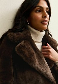 Bruine faux bont jas met brede kraag, gecombineerd met een crèmekleurige geribde coltrui. Soepele textuur, gedetailleerde stiksels en subtiele glans.