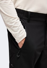 Main dans la poche d'un pantalon noir avec détail de poche zippée, portant un haut crème à manches longues.