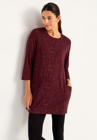 Kvinna lutad mot en vägg, klädd i en vinröd mönstrad tunika med 3/4-långa ärmar och framfickor, kombinerat med svarta leggings.