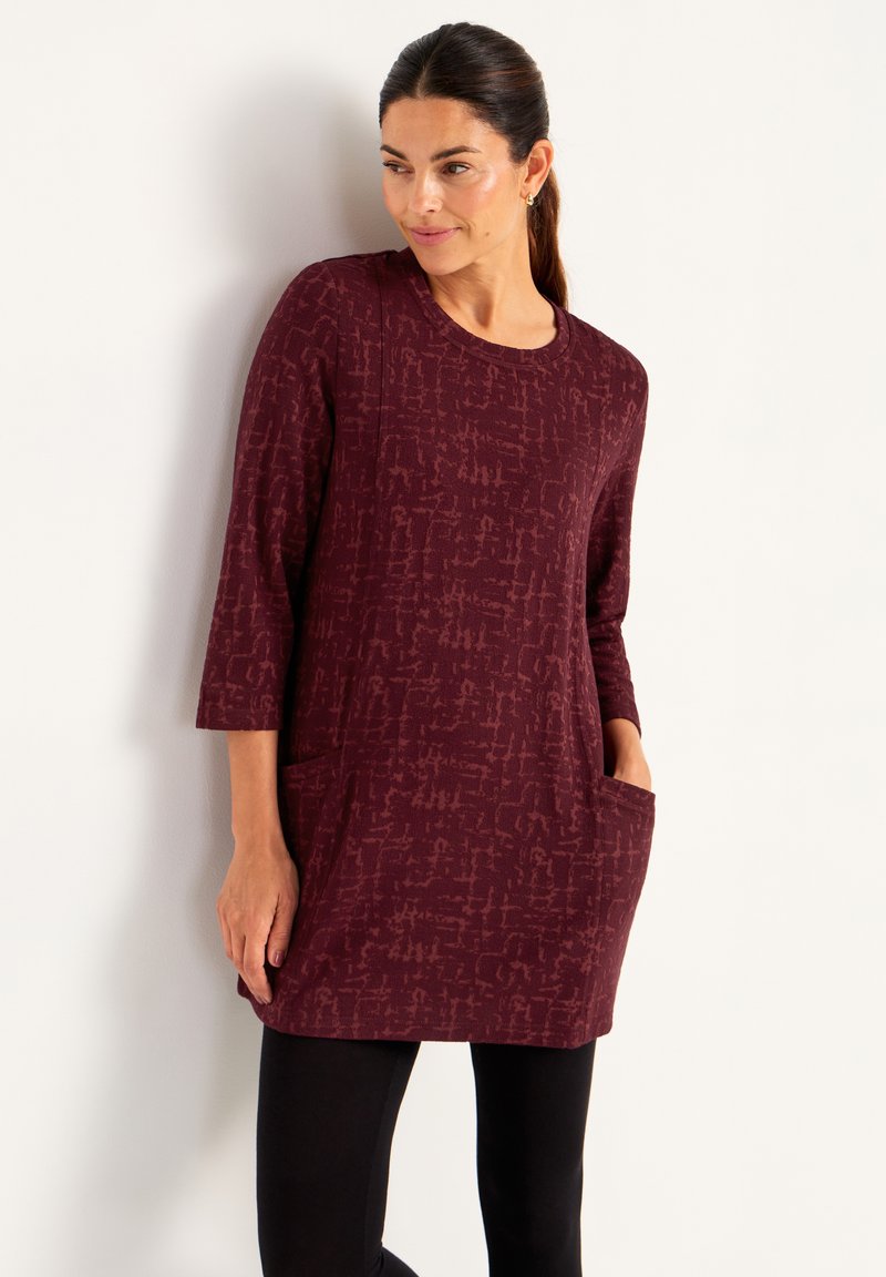 Kvinna lutad mot en vägg, klädd i en vinröd mönstrad tunika med 3/4-långa ärmar och framfickor, kombinerat med svarta leggings.