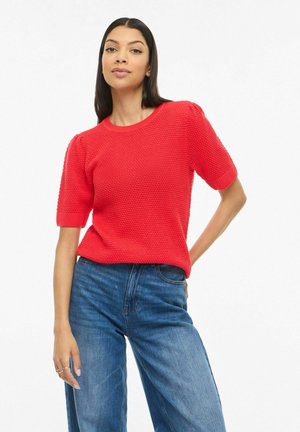 Frau trägt texturierten roten Kurzarmpullover und blaue hoch taillierte Jeans, posiert mit einer Hand auf der Hüfte vor weißem Hintergrund.