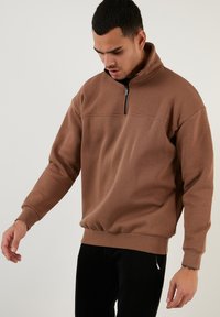 Sudadera pullover marrón con cremallera, cuello alto, puños acanalados y un ajuste relajado. Tejido suave con una textura lisa.
