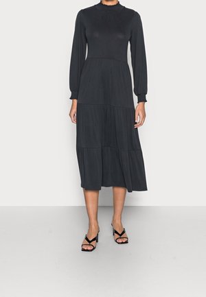 Schwarzes langärmliges Kleid aus glattem Stoff, mit hohem Kragen, gestuftem Design und gerafften Manschetten. Kombiniert mit schwarzen Riemchen-Highheels.